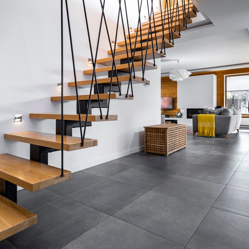 Escalier flottant en bois clair avec balustrade noire et éclairage mural intégré. Sol en carrelage gris. Salon moderne avec canapé gris et couverture jaune.