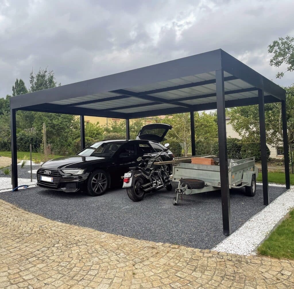 Abri voiture noir moderne protégeant une Audi break noire, une moto custom et une remorque sous un ciel nuageux. Allée pavée.