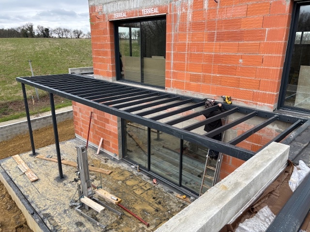 Construction d'une pergola métallique noire devant une maison en briques rouges, ouvrier travaillant près des baies vitrées. Le mot TERREAL est visible sur le mur.