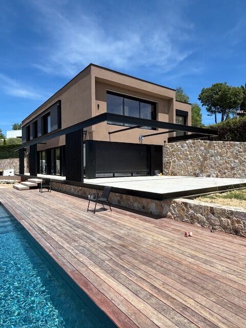 Villa moderne couleur terre cuite avec pergola noire, terrasse en bois longeant une piscine d'eau bleue, sous un ciel clair.